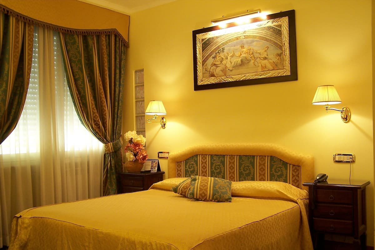 Una habitación de hotel con una cama grande, dos lámparas de noche, una mesita de noche, un jarrón con flores y un cuadro en la pared.