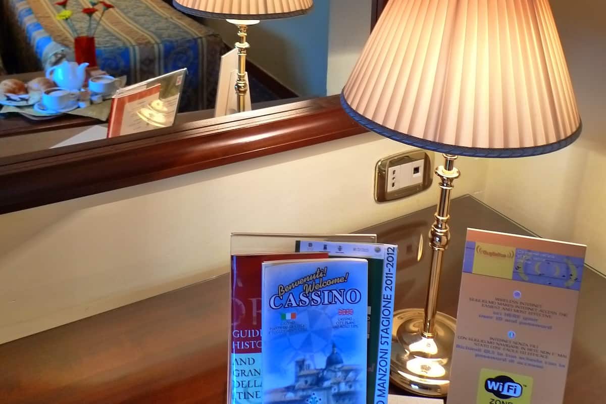 Habitación de hotel con cama, dos lámparas de noche, mesita de noche con un jarrón de flores, un cuadro enmarcado y un cartel de bienvenida.