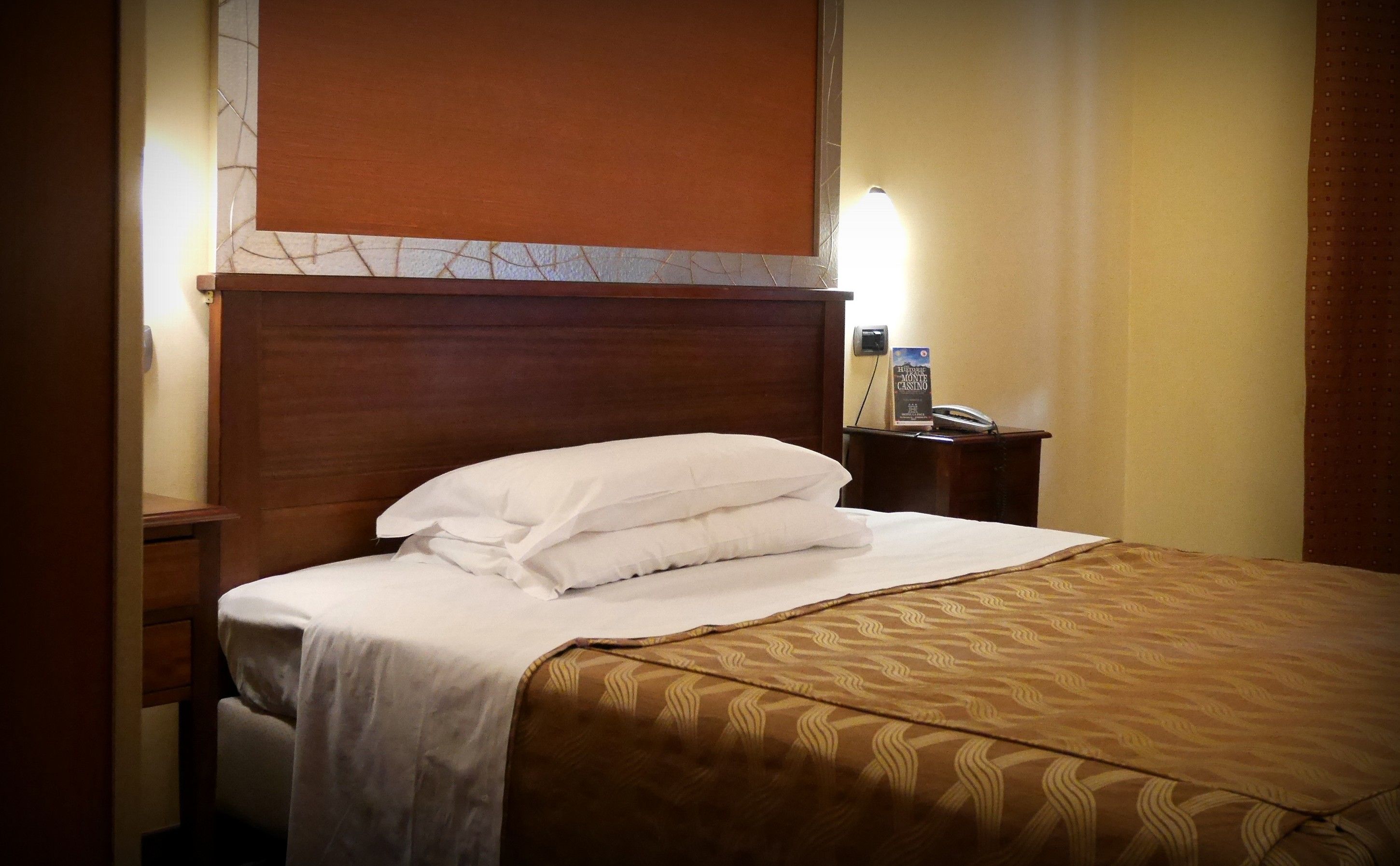 Una habitación de hotel con cabecera de madera, cama con sábanas blancas y cubrecama marrón estampado, mesita de noche con teléfono y cartel, y pared con diseño que imita mármol.