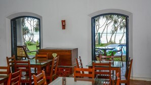 A dining area with wooden chairs and tables, a central wooden cabinet, and large arched windows with a view of palm trees and a pool.