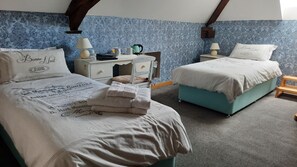 Un dormitorio con dos camas, cada una con cubrecama y almohadas blancas a juego. Hay mesitas de noche con lámparas y un pequeño estante. Las paredes tienen un papel tapiz azul con estampado.