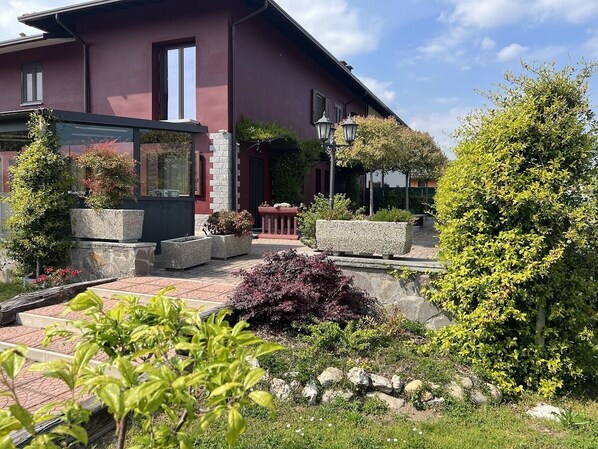 A modern building with a red facade, stone planters, and a well-maintained garden with various shrubs and a tree. - Villa Rosy B&B (Paruzzaro)