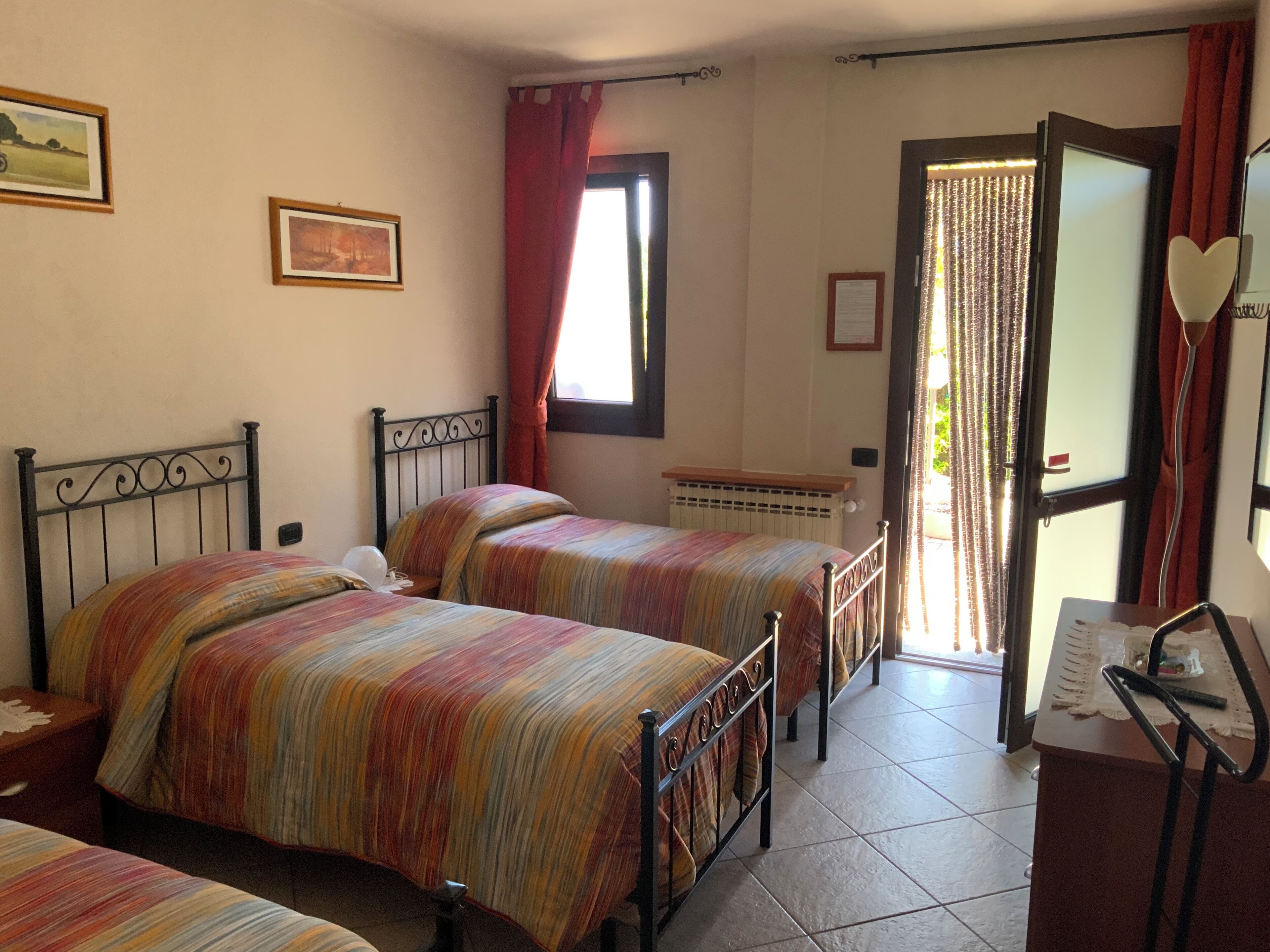 A bedroom with two beds, a radiator, a window with curtains, and a door leading outside.