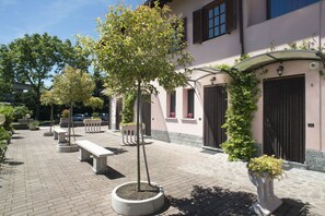 A courtyard with benches, potted plants, and a building with a pink facade and black shutters. - Villa Rosy B&B (Paruzzaro)