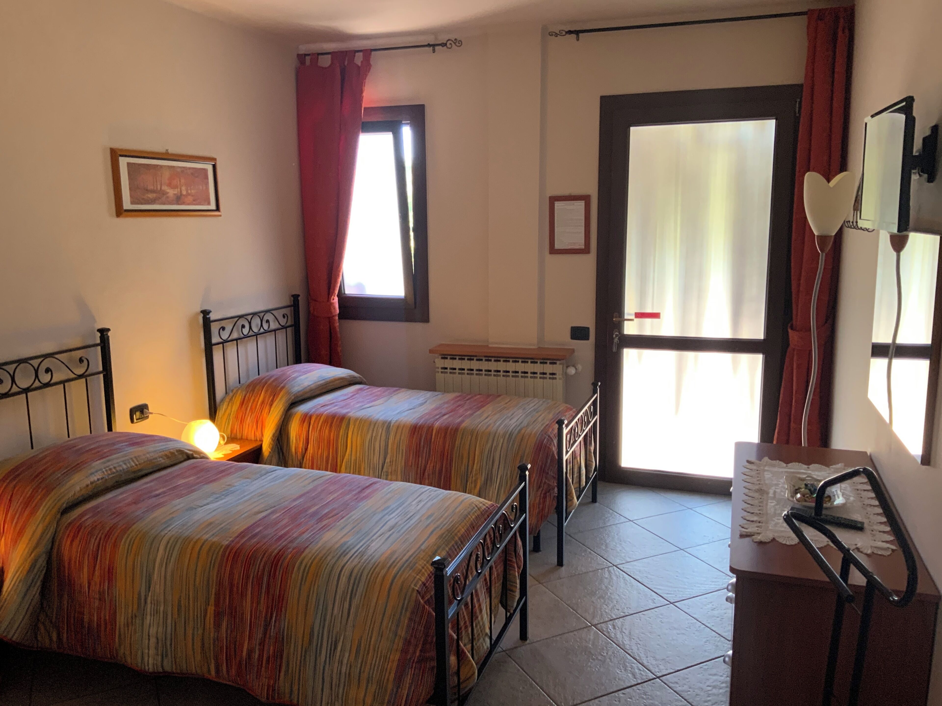 A room with two beds, a radiator, a desk with a chair, a window with curtains, and a framed picture on the wall.