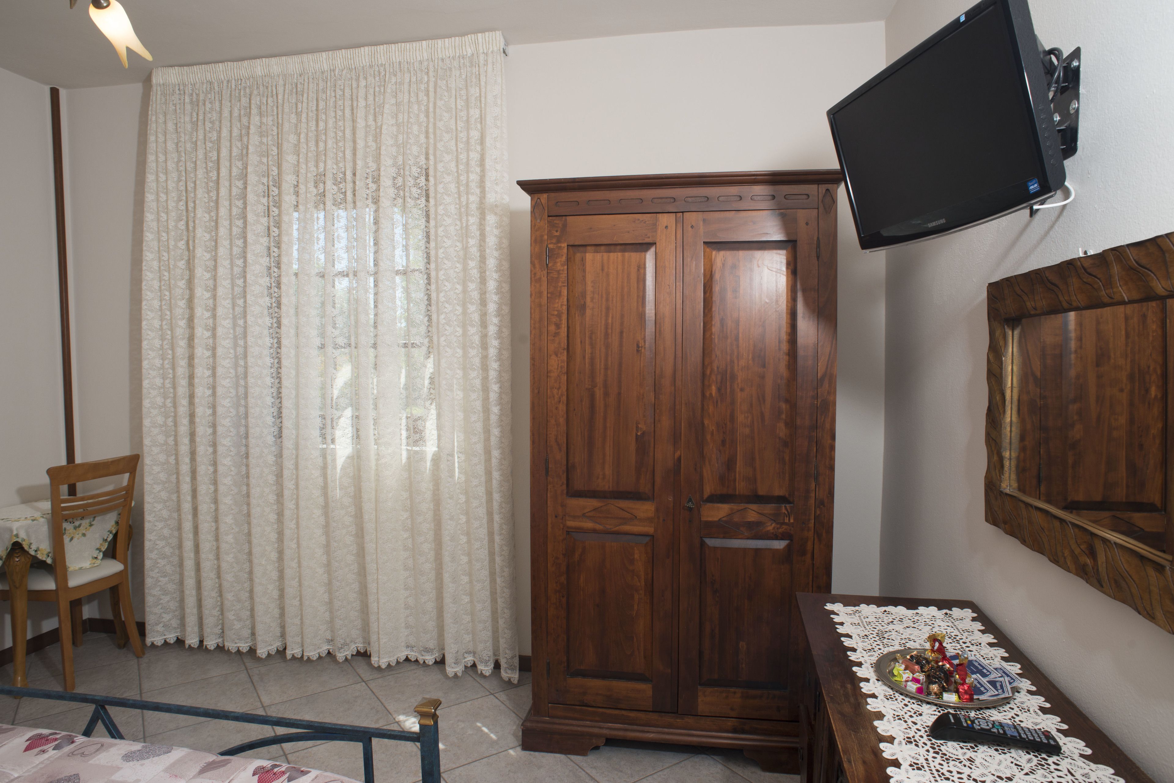A room with a wooden wardrobe, a television mounted on the wall, a table with a lace tablecloth, and a chair.
