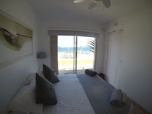 A bedroom with a bed, pillows, a bedside lamp, a view of the beach, and a ceiling fan.