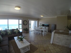 Ampla sala de estar com sofá, mesa de centro e mesa de jantar. Há plantas em vasos e vista para o oceano por grandes janelas.