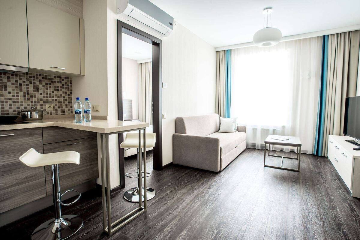 Ein modernes Hotelzimmer mit einer Küchenzeile, einem Essbereich, einem Sofa, einem Fernseher und einem großen Fenster mit Vorhängen.