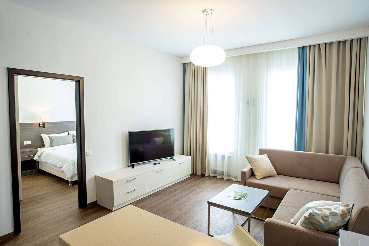 Ein modernes Hotelzimmer mit einem Bett, einem Fernseher auf einem weißen Schrank, einer Couch, einem Couchtisch und einem großen Spiegel.