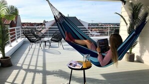 Eine Person entspannt in einer Hängematte auf einem Balkon mit Tisch, Stühlen und Blumentöpfen.