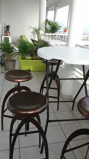 Une terrasse avec une table blanche et quatre tabourets, entourée de plantes en pot et d’une rambarde blanche.
