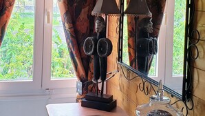 Et værelse med et stort vindue, et spejl med dekorativ ramme, en lampe og et lille bord med en notesbog og et glas.