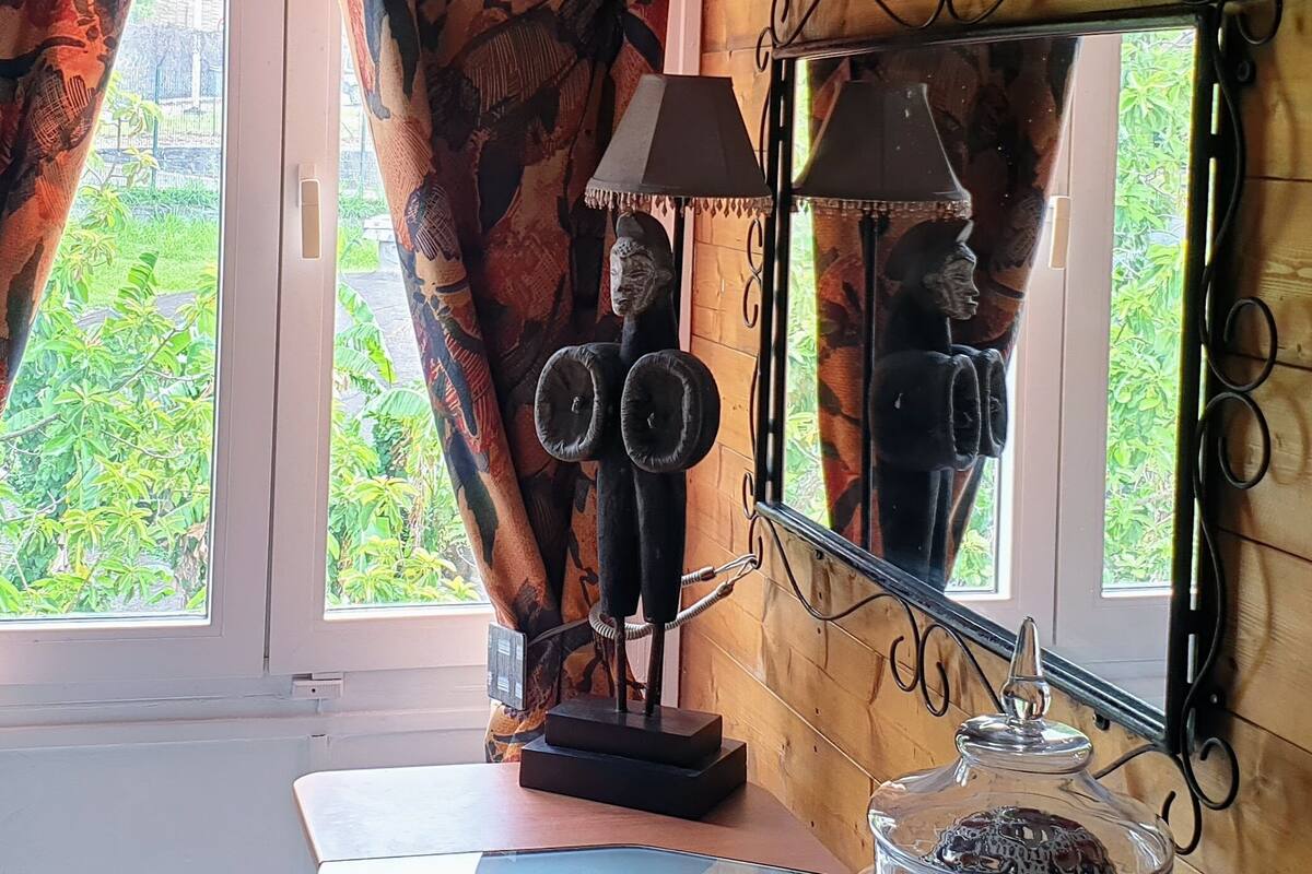 A room with a large window, a mirror with a decorative frame, a lamp, and a small table with a notebook and a glass jar.