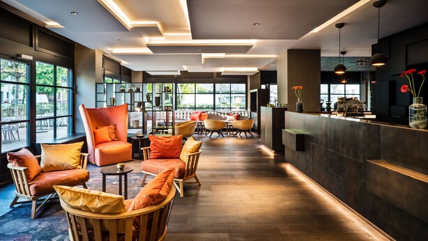 Un moderno vestíbulo de hotel con suelo de madera, un mostrador de recepción y áreas de descanso con sillas anaranjadas y de madera.