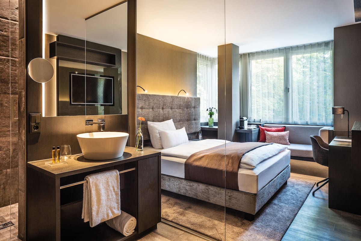 A modern hotel room with a large bed, a built-in vanity with a large sink, a flat-screen TV, and a large window providing a view of the outdoors.