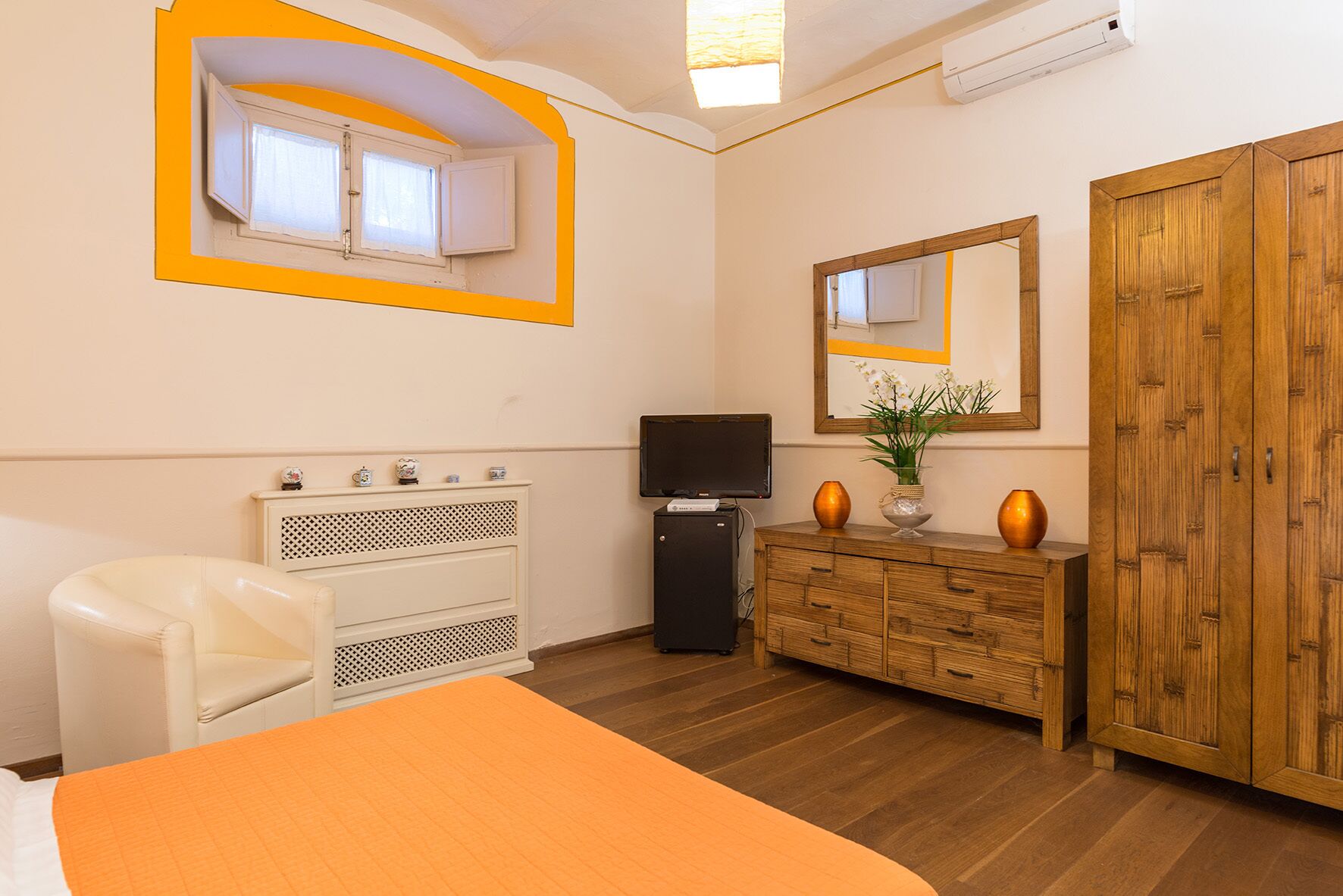 Une chambre à coucher comprenant un lit, une commode, un téléviseur, une armoire et une fenêtre avec des rideaux.
