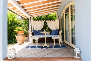 A covered patio with a cushioned seating area, a table, and a large potted plant.