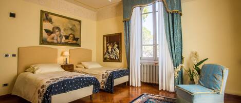 Quarto de hotel com duas camas, um candelabro, um tapete azul estampado e uma cadeira.
