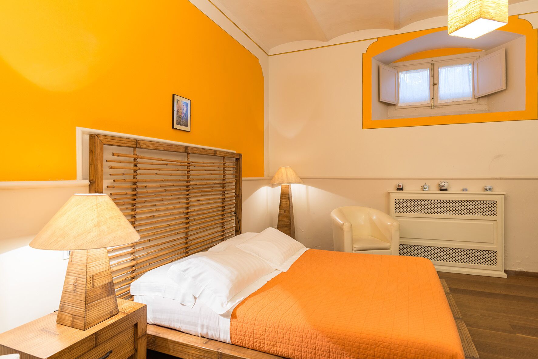 Et soveværelse med sengegavl i træ, seng med hvidt og orange sengetøj, et natbord med lampe, en stol og et lille skab med pynteting.
