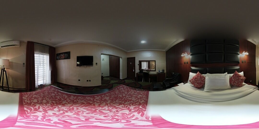 A panoramic view of a hotel room with a bed, a desk, a chair, a television, and a window with curtains.