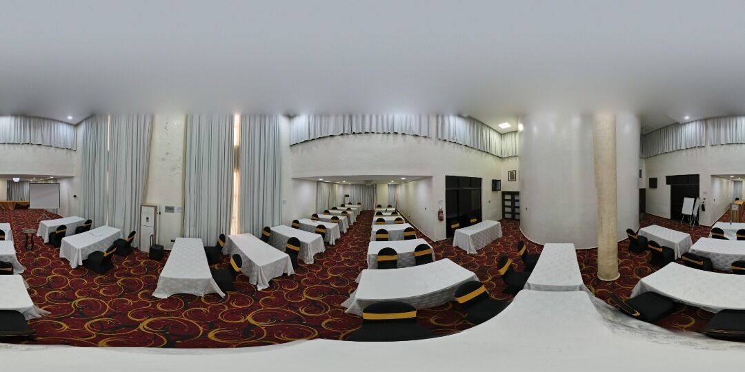 Uma sala de conferências com mesas e cadeiras dispostas em fileiras, um tapete vermelho e cortinas brancas.
