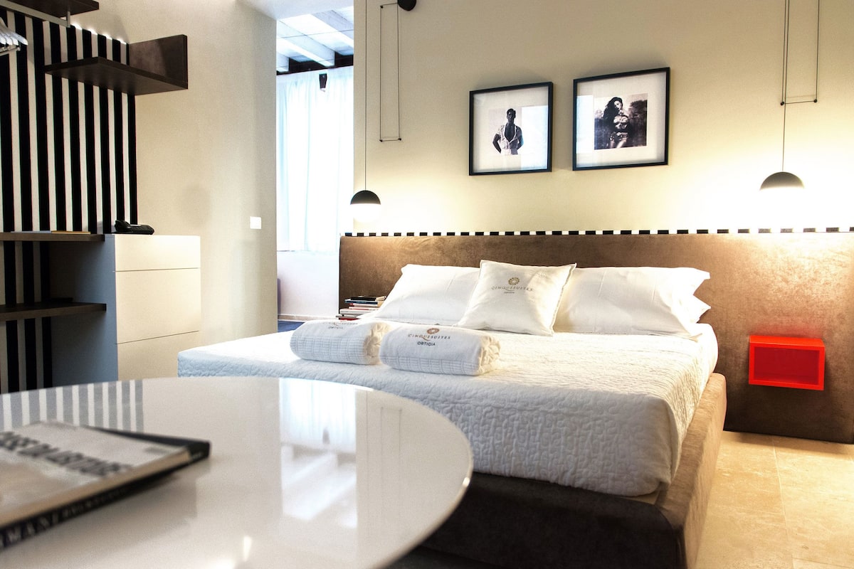 A neatly made bed with white linens and pillows, a bedside table with a book, and a red object on the nightstand.
