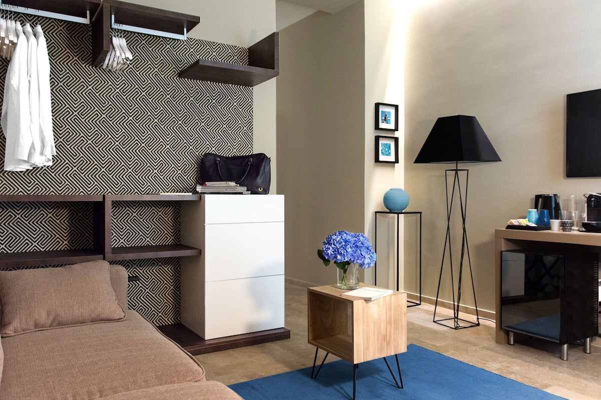 A modern hotel room with a sofa, a small wooden side table with a vase of flowers, a desk with a lamp, and a wardrobe with hanging clothes.