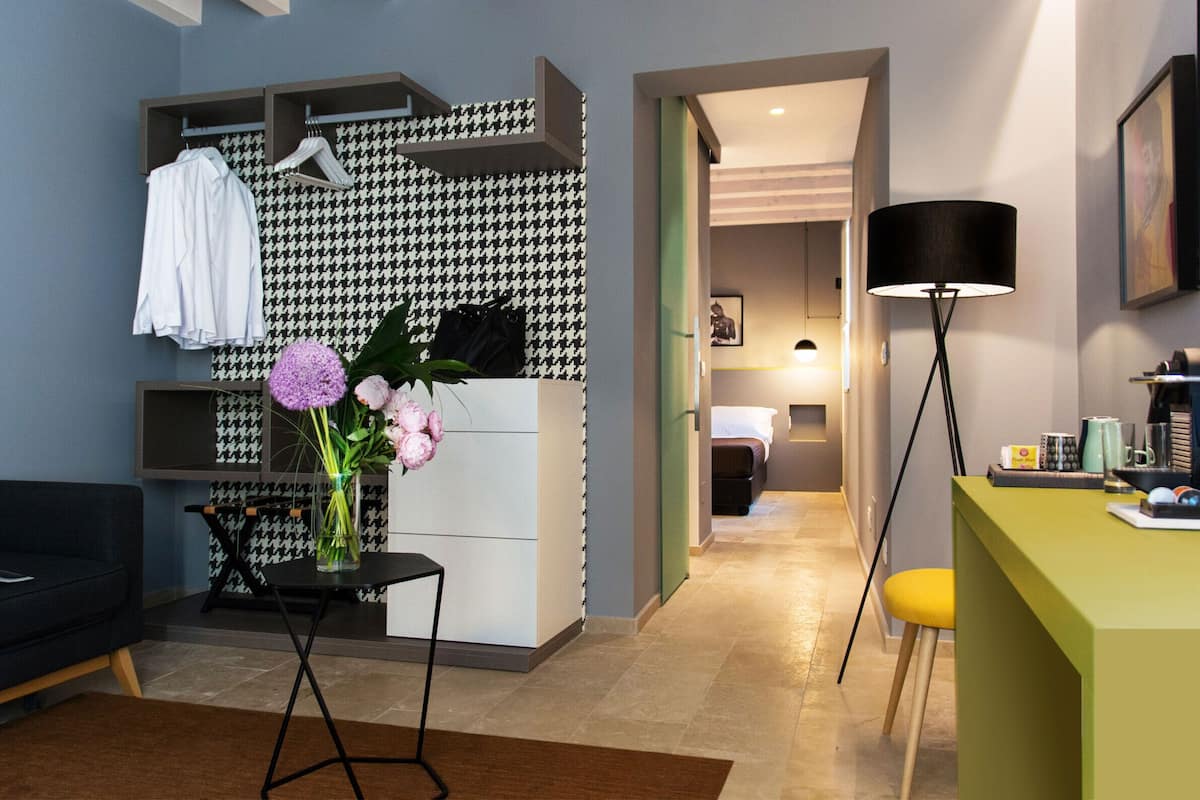 A modern hotel room with a sofa, a small table with a vase of flowers, a desk with a lamp, and a houndstooth-patterned wall.