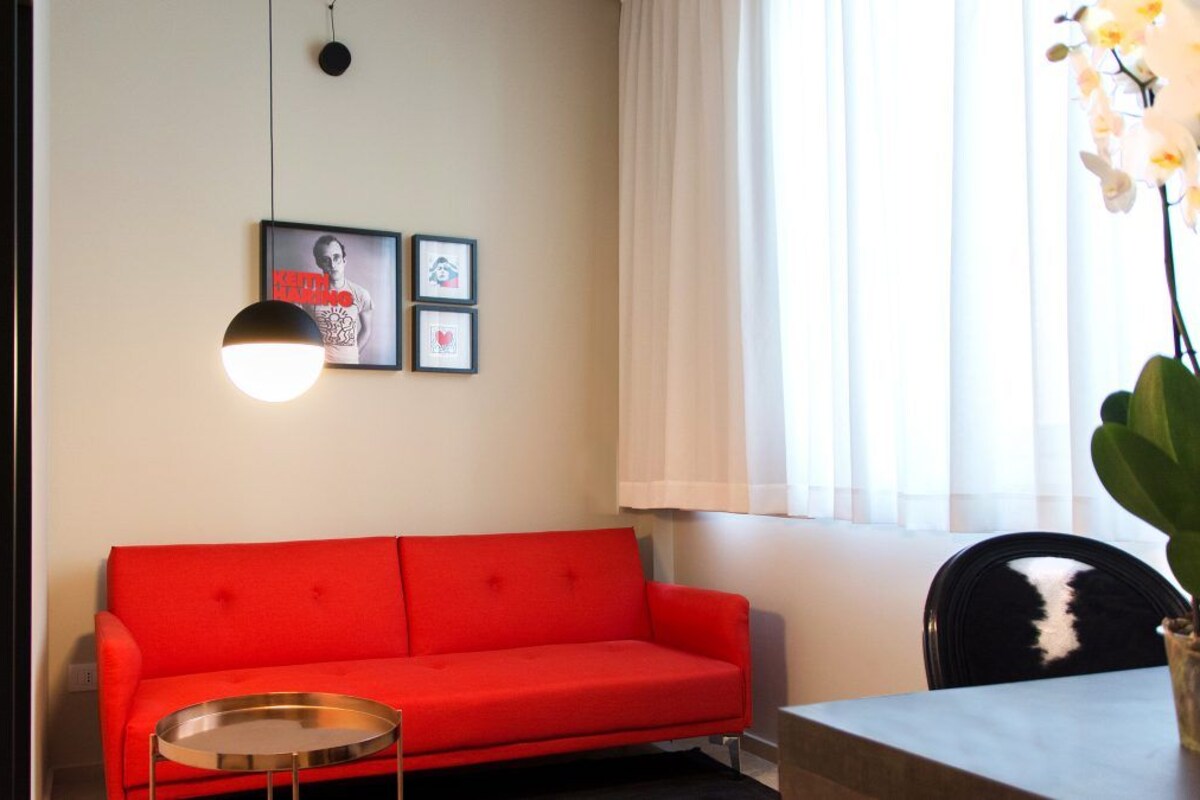 A red sofa, a round table, and a chair in a room with a white wall, a hanging light, and a window with curtains.