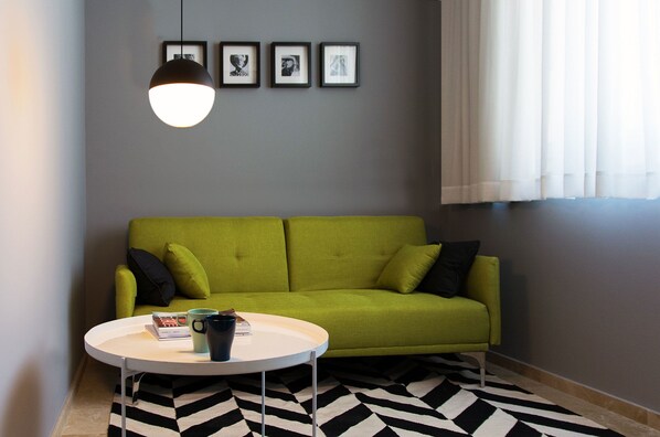 Un sofá verde con cojines negros, una mesa redonda blanca y una alfombra blanco y negro, con una lámpara colgante.