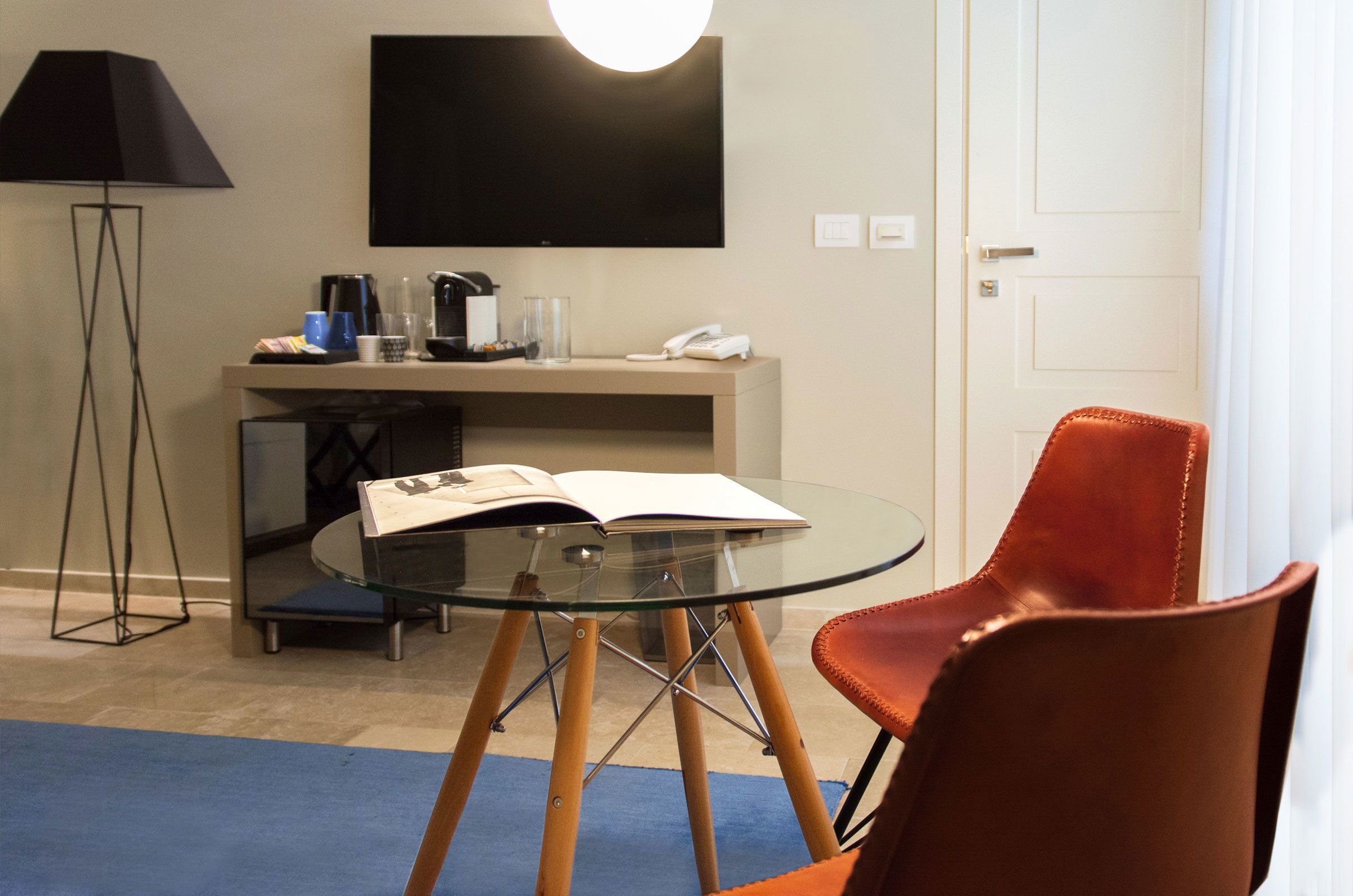 A hotel room with a glass table, a red chair, a lamp, a flat-screen TV, a coffee maker, and a telephone.