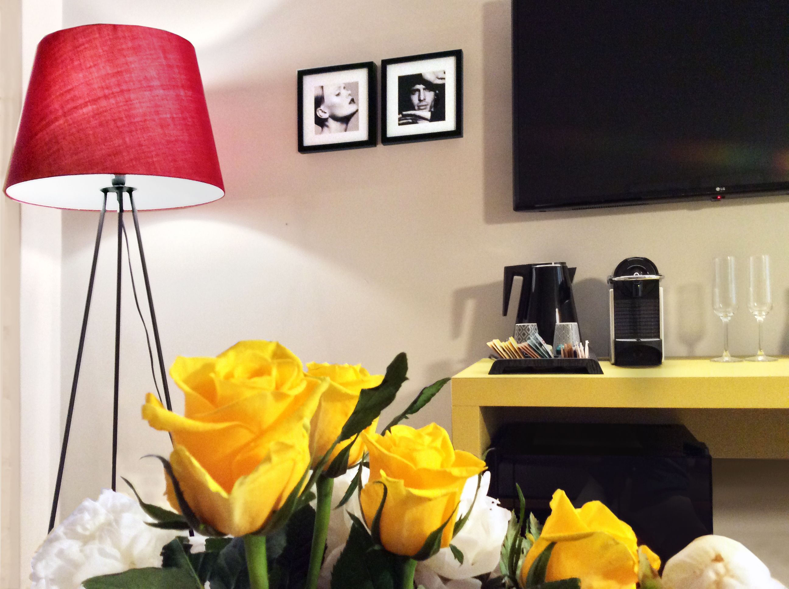 A room with a red floor lamp, a yellow side table with a coffee maker and glasses, and a bouquet of yellow and white flowers.