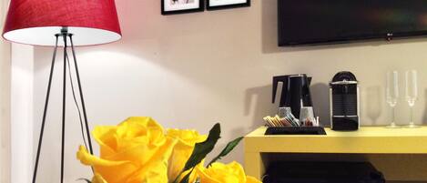 A room with a red floor lamp, a yellow side table with a coffee maker and glasses, and a bouquet of yellow and white flowers.