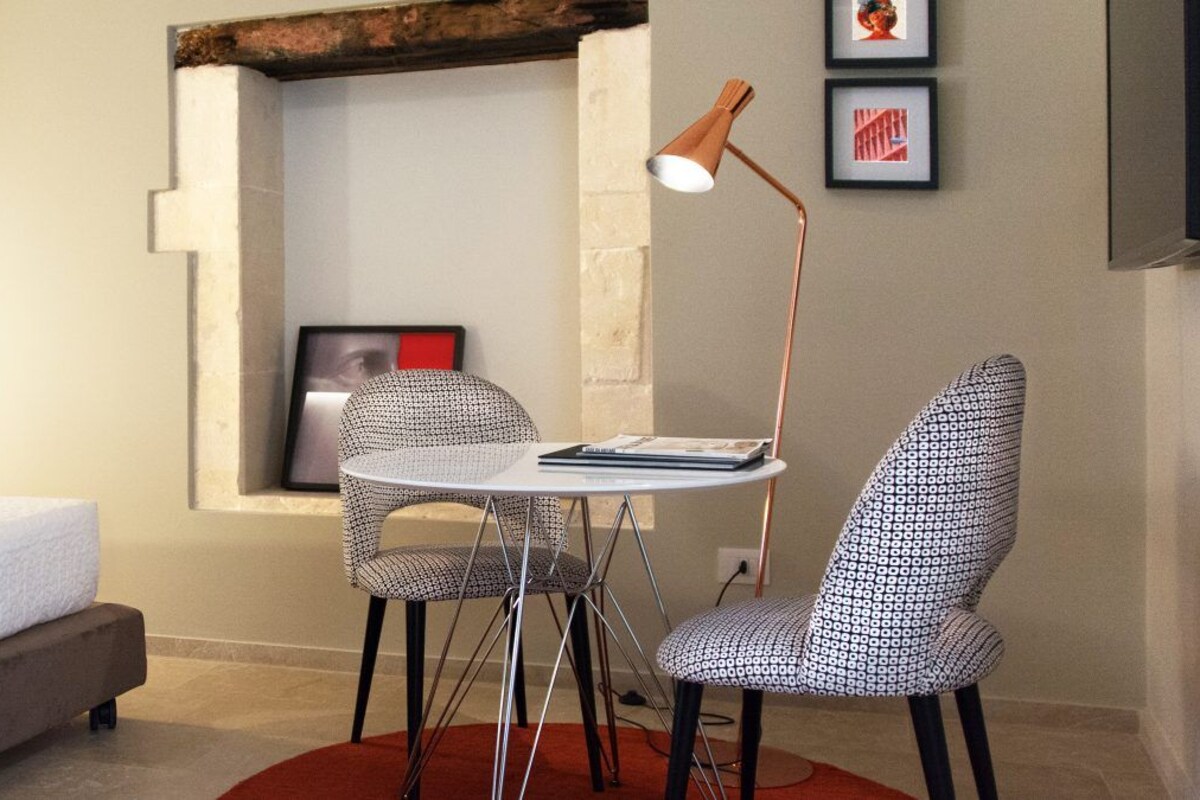 A small room with a bed, a round table with two patterned chairs, a floor lamp, and a framed picture on the wall.
