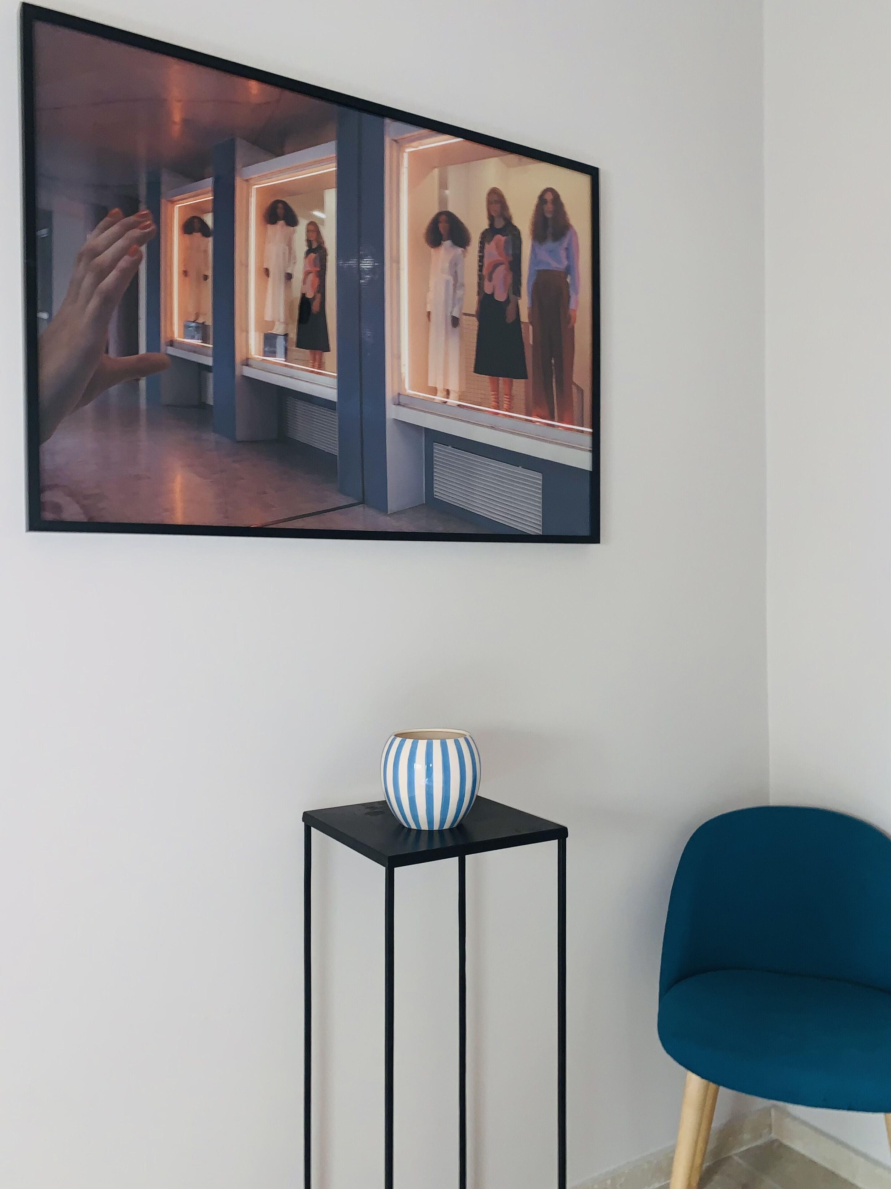 A framed photograph of a store window with mannequins, a small table with a striped vase, and a blue chair.