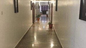 A long, narrow hallway with a polished floor, potted plants, and framed pictures on the walls. - The Den Hotel (Abuja)