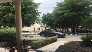 A covered outdoor area with a paved walkway, potted plants, and parked cars. - The Den Hotel (Abuja)