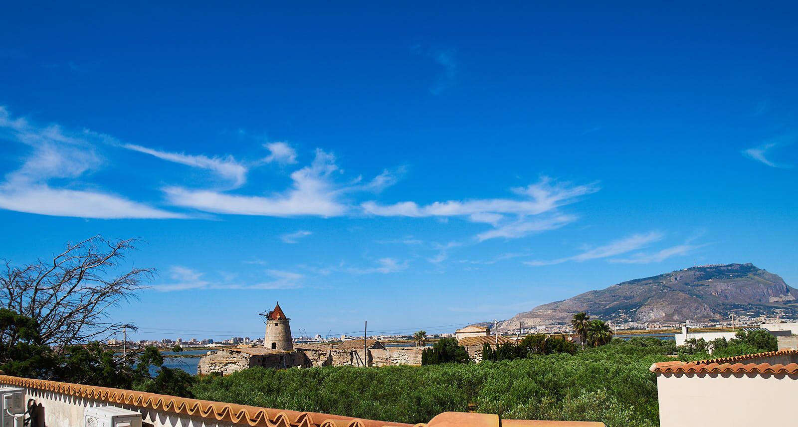 Un pueblo costero con un molino de viento destacado, una montaña al fondo y un cielo azul despejado.