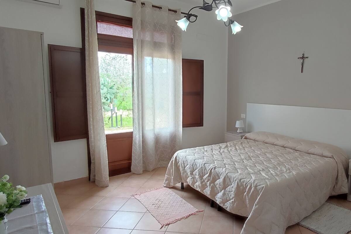 Ein Schlafzimmer mit Bett, Nachttischen, einer Lampe, einem Kreuz an der Wand und einem Fenster mit Vorhängen.
