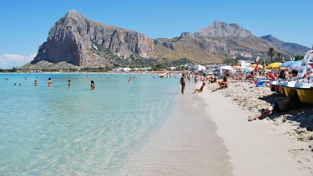 Ein Strand mit klarem türkisblauem Wasser, Menschen beim Schwimmen und Sonnenbaden, und Berge im Hintergrund.
