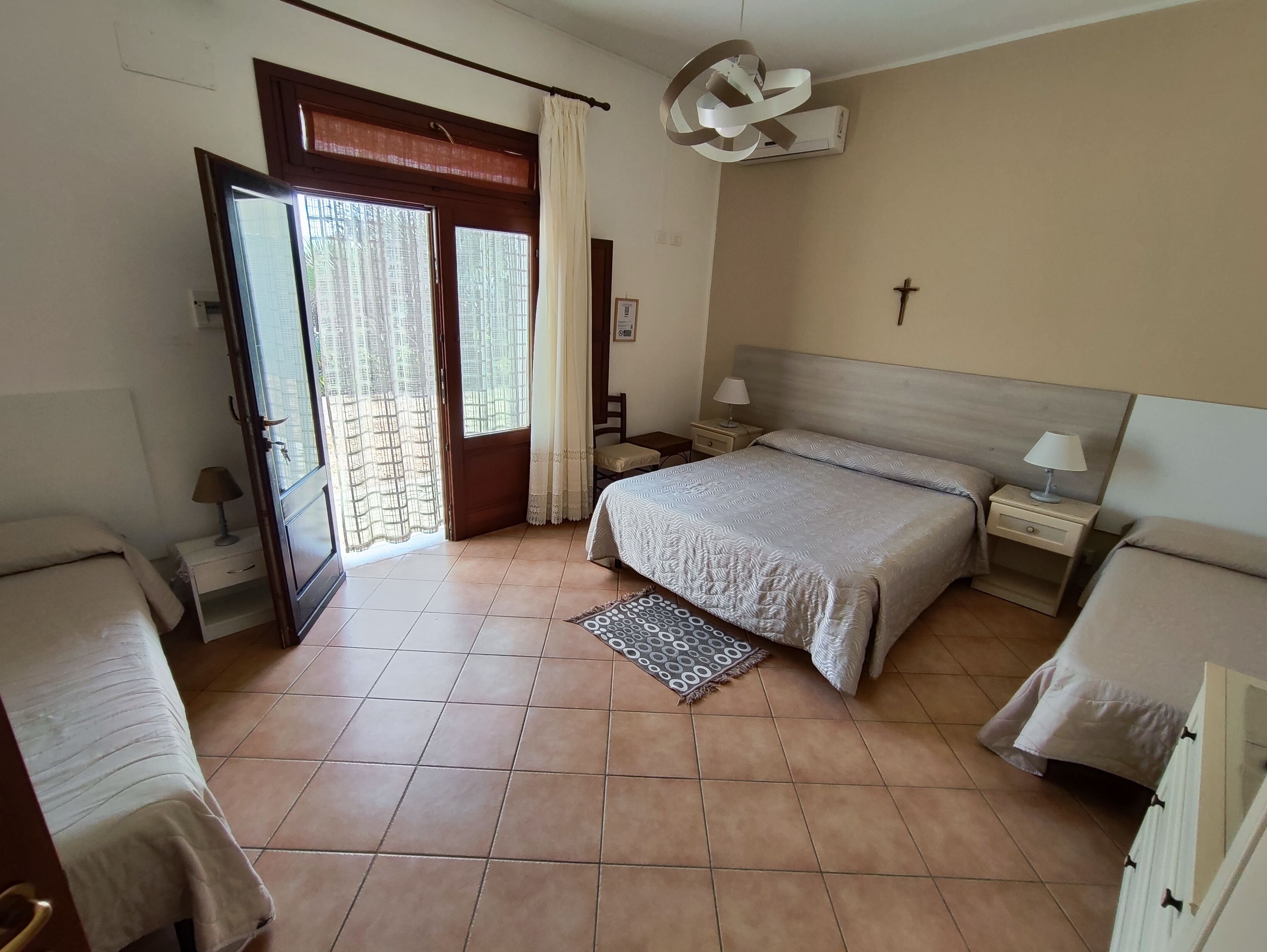 Ein Schlafzimmer mit einem Bett, zwei Nachttischen, einem Fenster mit Vorhängen, einem Deckenventilator und einem Kreuz an der Wand.