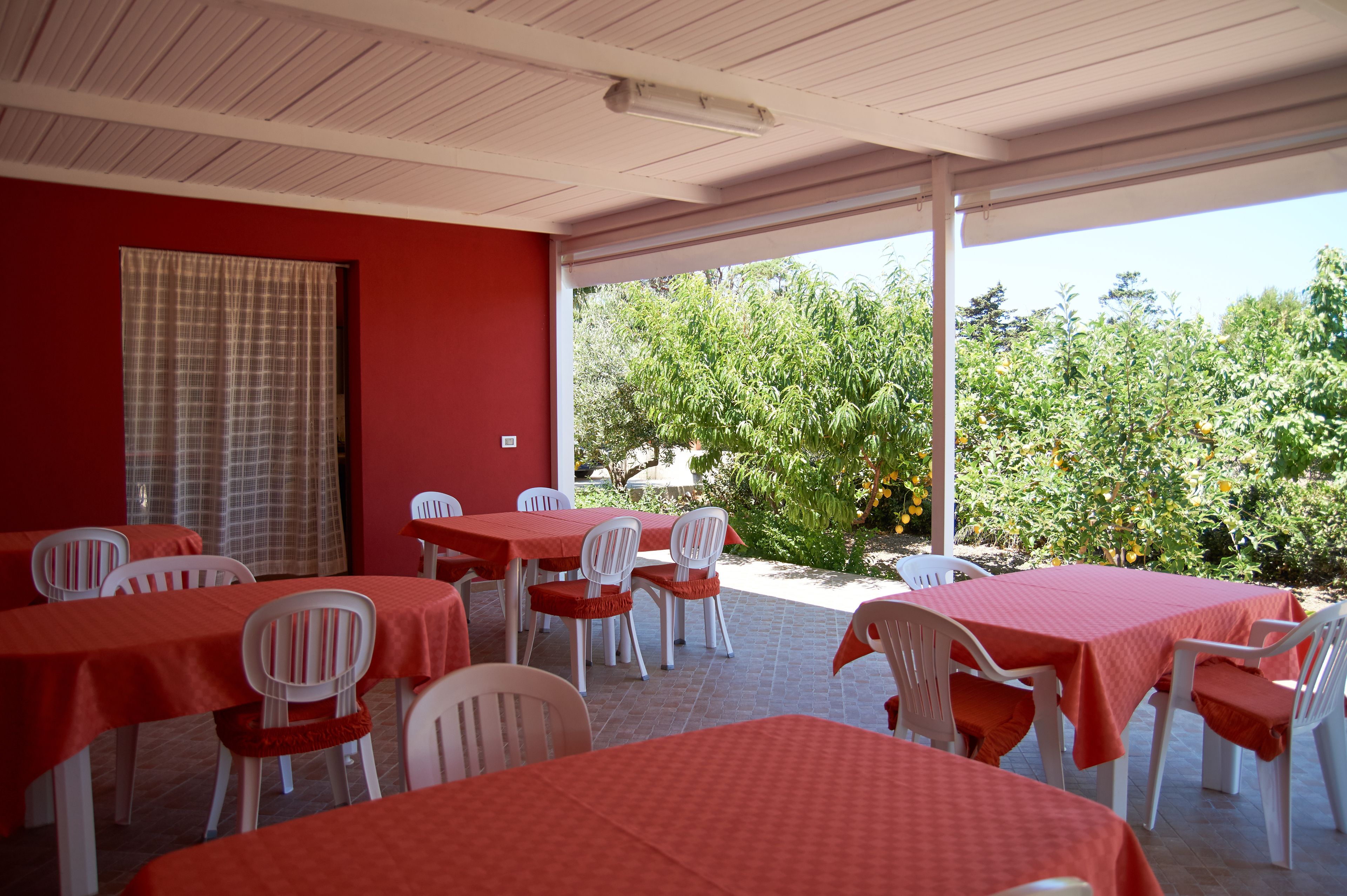 An outdoor patio area with tables and chairs, covered by a roof, and a view of trees and plants.