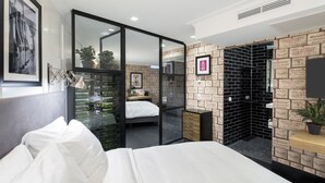 Una habitación de hotel moderna con cama, escritorio y un espejo grande. Se observa una pared con diseño de ladrillo y una pared de ladrillo negro con texto.
