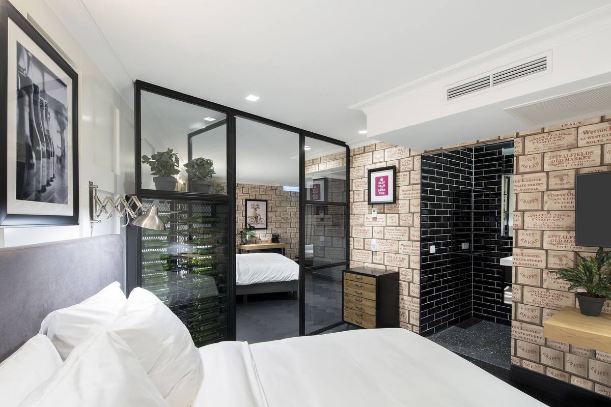 A modern hotel room with a bed, a desk, and a large mirror. There is a brick-patterned wall and a black brick wall with text.