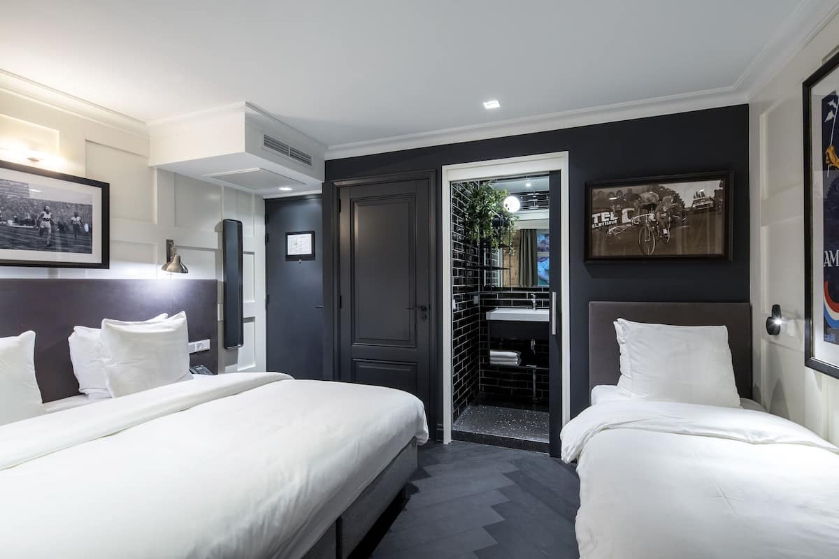 A hotel room with two beds, a dark accent wall, a framed picture, a mirror, and a door leading to a bathroom.