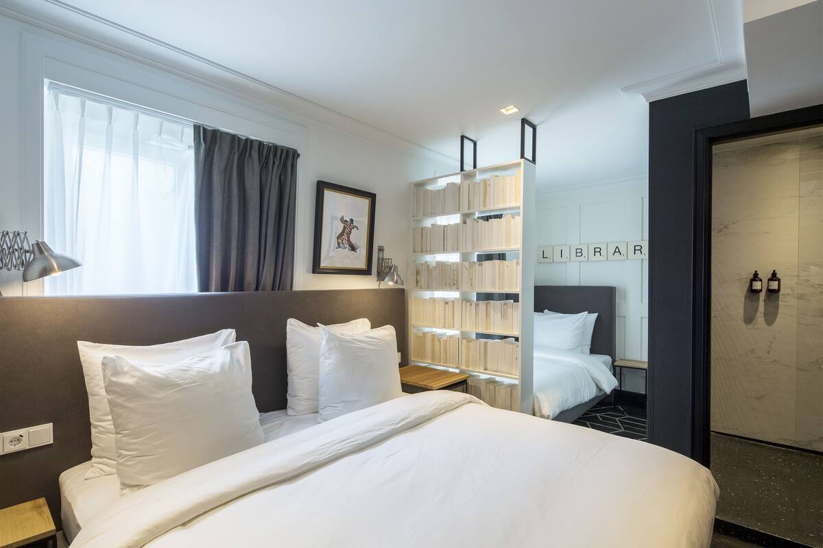 A modern hotel room with a large bed, a bedside table, a bookshelf, and a view of the city.