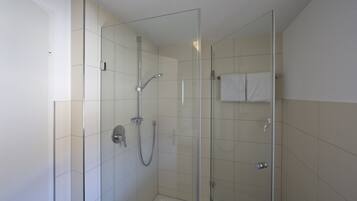 A glass-enclosed shower with a handheld showerhead and a fixed showerhead, white tiled walls, and a towel hanging on the wall.