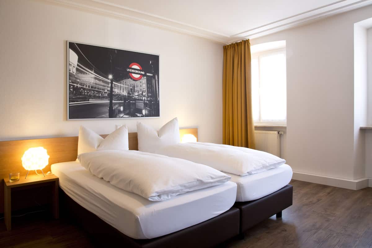 A hotel room with a double bed, a nightstand with a lamp, a framed picture of the London Underground, and a window with curtains.