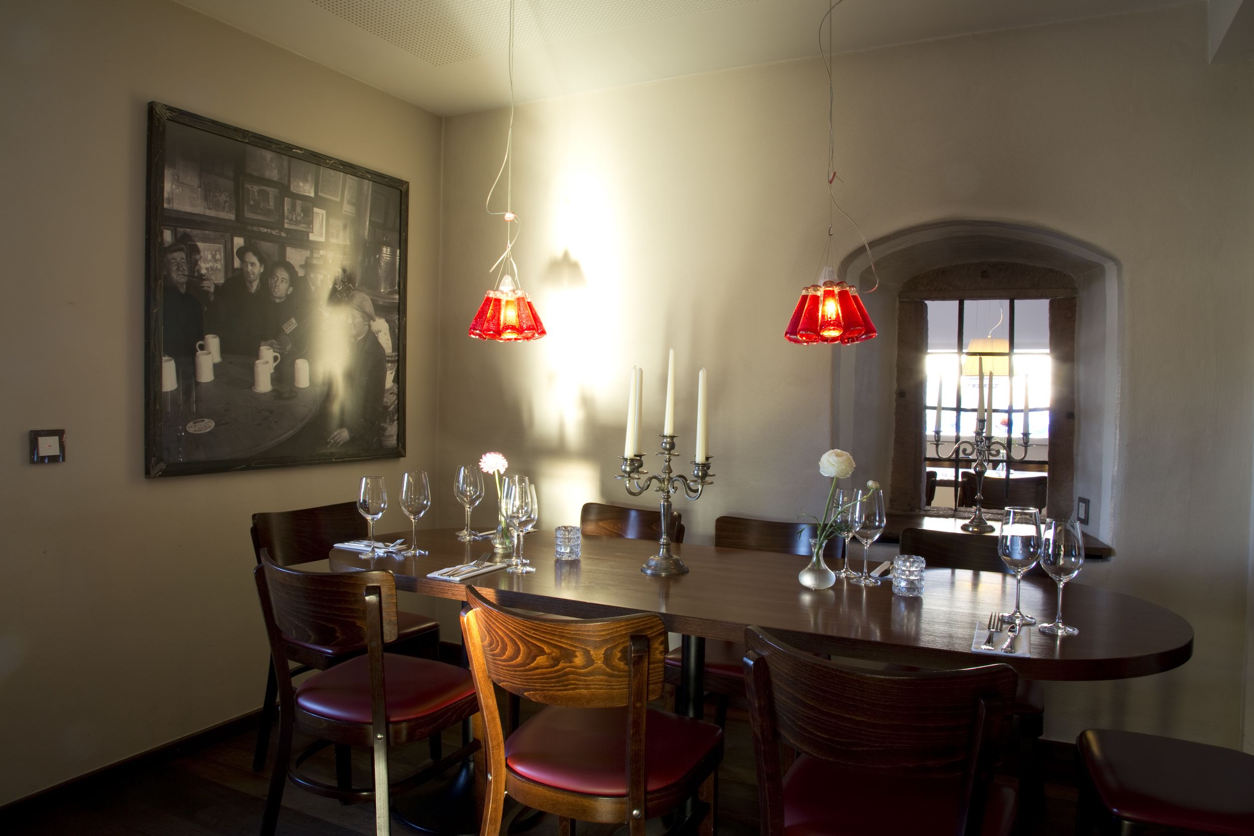 Un comedor con una mesa de madera dispuesta para seis personas, rodeada de sillas de madera con cojines rojos. Dos lámparas colgantes rojas se encuentran sobre la mesa.